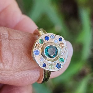 K&ouml;nnte beinhalten: Ein goldener Ring mit einer runden, strukturierten Fassung, in der ein gr&uuml;ner Edelstein von einem Heiligenschein aus kleineren Edelsteinen in verschiedenen Farben, darunter Blau, Gr&uuml;n, Wei&szlig; und Gelb, umgeben ist.