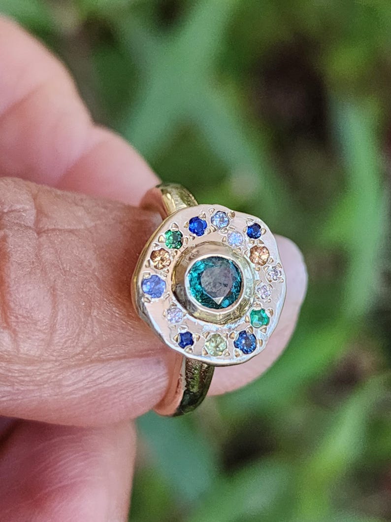 K&ouml;nnte beinhalten: Ein goldener Ring mit einer runden, strukturierten Fassung, in der ein gr&uuml;ner Edelstein von einem Heiligenschein aus kleineren Edelsteinen in verschiedenen Farben, darunter Blau, Gr&uuml;n, Wei&szlig; und Gelb, umgeben ist.