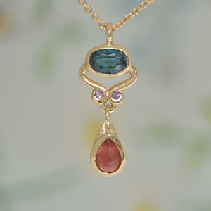May include: A gold pendant necklace with a teardrop-shaped red gemstone and a blue gemstone set in a gold frame with two small purple gemstones.