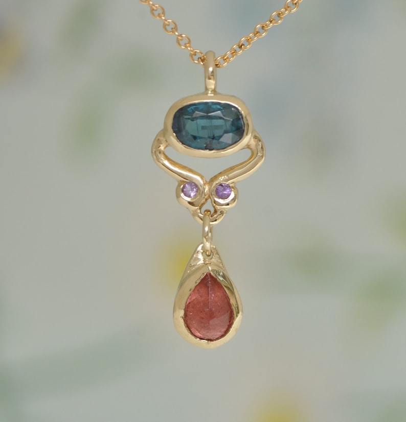 May include: A gold pendant necklace with a teardrop-shaped red gemstone and a blue gemstone set in a gold frame with two small purple gemstones.