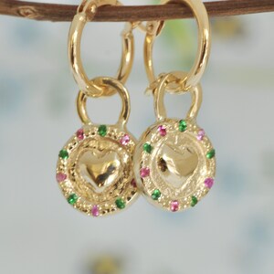May include: Gold hoop earrings with a heart-shaped charm. The charm is surrounded by a circle of pink and green gemstones.