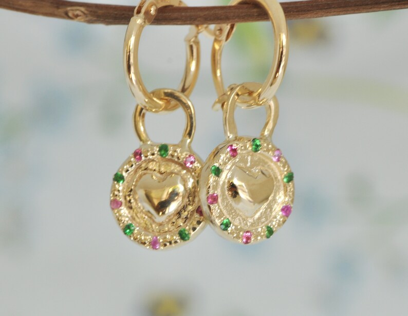 May include: Gold hoop earrings with a heart-shaped charm. The charm is surrounded by a circle of pink and green gemstones.