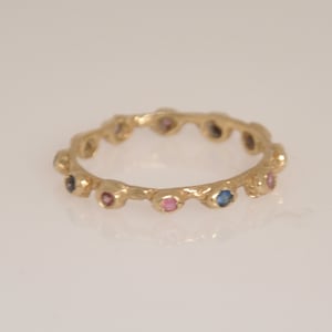 May include: A gold band ring with a textured surface and a row of small, colorful gemstones. The gemstones are blue, pink, and clear.