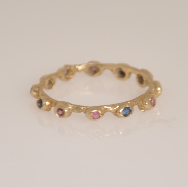 May include: A gold band ring with a textured surface and a row of small, colorful gemstones. The gemstones are blue, pink, and clear.