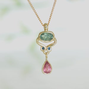 May include: A gold necklace with a pendant featuring a teardrop-shaped pink gemstone and a larger oval-shaped green gemstone surrounded by two smaller blue gemstones.