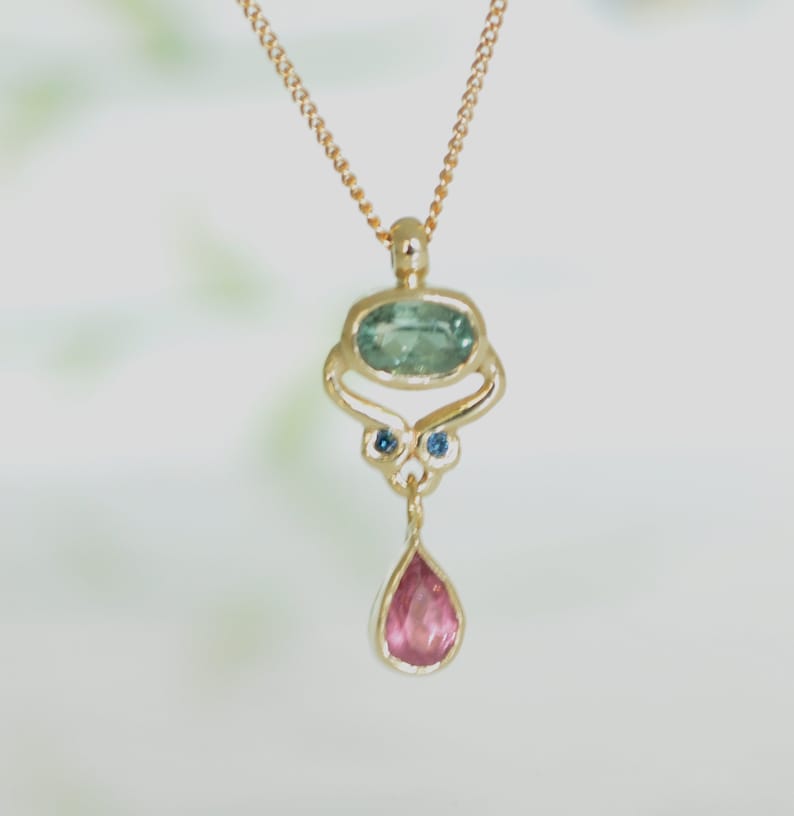 May include: A gold necklace with a pendant featuring a teardrop-shaped pink gemstone and a larger oval-shaped green gemstone surrounded by two smaller blue gemstones.