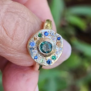 K&ouml;nnte beinhalten: Ein goldener Ring mit einem gr&uuml;nen Edelstein, der von einem Heiligenschein aus kleineren Edelsteinen in verschiedenen Farben, darunter Blau, Wei&szlig; und Gelb, umgeben ist.