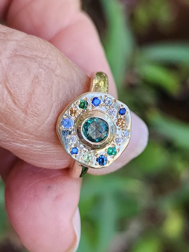 K&ouml;nnte beinhalten: Ein goldener Ring mit einem gr&uuml;nen Edelstein, der von einem Heiligenschein aus kleineren Edelsteinen in verschiedenen Farben, darunter Blau, Wei&szlig; und Gelb, umgeben ist.