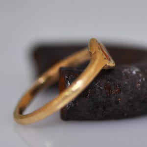 May include: A gold ring with a small, oval-shaped orange stone. The ring is set on a dark, textured surface, with the band curving away from the stone. The ring appears to be vintage or antique.
