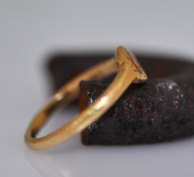 May include: A gold ring with a small, oval-shaped orange stone. The ring is set on a dark, textured surface, with the band curving away from the stone. The ring appears to be vintage or antique.