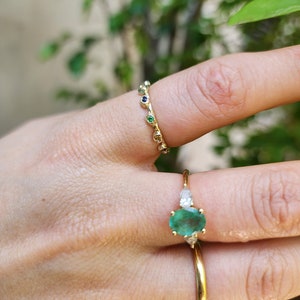 May include: Two gold rings with gemstones. The first ring has a large oval emerald gemstone with two smaller diamond gemstones on either side. The second ring has a band of small gemstones in various colors, including green, blue, and white.