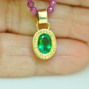 Pu&ograve; includere: Un pendente in oro con una gemma ovale verde smeraldo, circondata da un bordo dorato decorativo. Il pendente &egrave; appeso a una collana di piccole perle rosa rotonde. Il gioiello &egrave; mostrato su uno sfondo sfocato.