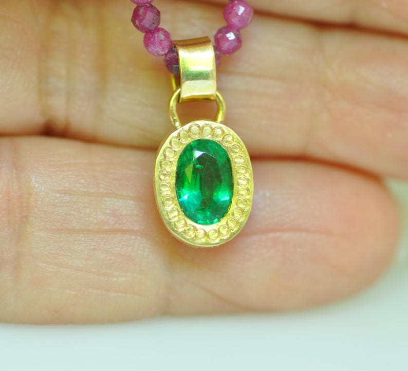 Pu&ograve; includere: Un pendente in oro con una gemma ovale verde smeraldo, circondata da un bordo dorato decorativo. Il pendente &egrave; appeso a una collana di piccole perle rosa rotonde. Il gioiello &egrave; mostrato su uno sfondo sfocato.