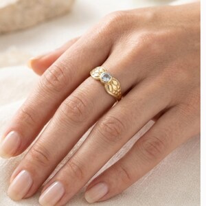 May include: A gold ring with a floral design and a small, light blue gemstone. The ring is on a hand with manicured nails, resting on a white surface. The ring's intricate details and the gemstone are the focal points.