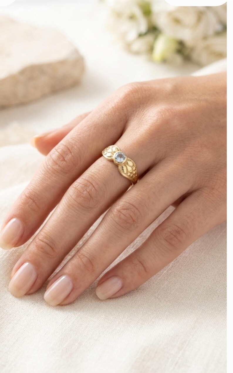 May include: A gold ring with a floral design and a small, light blue gemstone. The ring is on a hand with manicured nails, resting on a white surface. The ring's intricate details and the gemstone are the focal points.