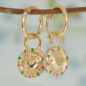 May include: Gold hoop earrings with a heart-shaped charm. The charm is decorated with a green and pink gemstone border.