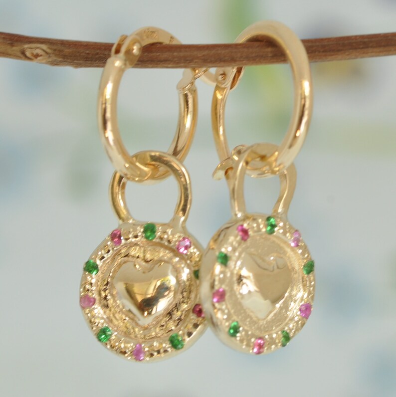 May include: Gold hoop earrings with a heart-shaped charm. The charm is decorated with a green and pink gemstone border.