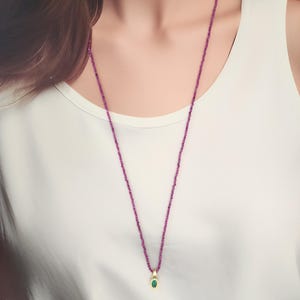 Pu&ograve; includere: Una lunga e delicata collana con una fila di piccole perline rosa. La collana presenta un ciondolo dorato con una piccola pietra verde di forma ovale. La collana &egrave; indossata sopra un top bianco.