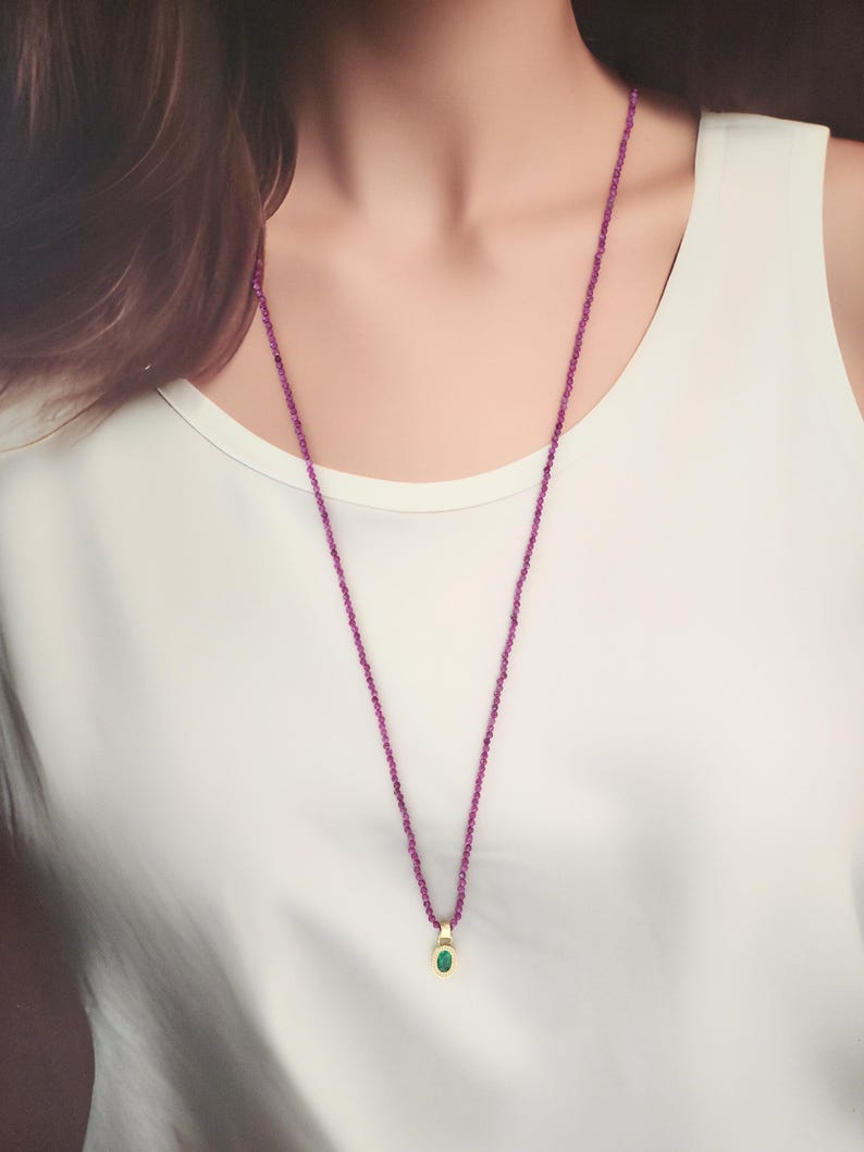 Pu&ograve; includere: Una lunga e delicata collana con una fila di piccole perline rosa. La collana presenta un ciondolo dorato con una piccola pietra verde di forma ovale. La collana &egrave; indossata sopra un top bianco.