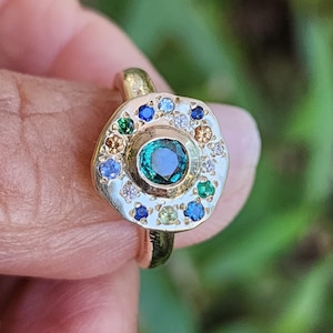 K&ouml;nnte beinhalten: Ein goldener Ring mit einem gr&uuml;nen Edelstein, der von kleineren Edelsteinen in verschiedenen Farben, darunter Blau, Gelb und Wei&szlig;, umgeben ist.