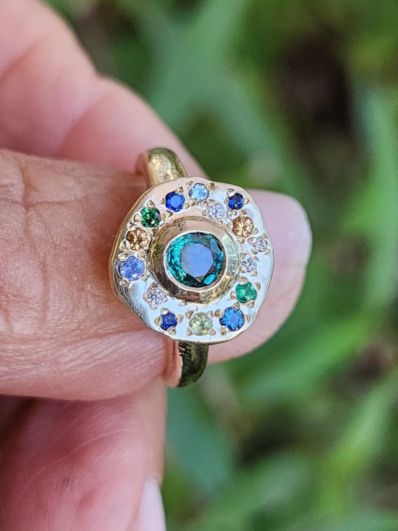 K&ouml;nnte beinhalten: Ein goldener Ring mit einem gr&uuml;nen Edelstein, der von kleineren Edelsteinen in verschiedenen Farben, darunter Blau, Gelb und Wei&szlig;, umgeben ist.