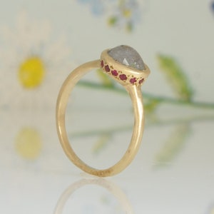 Op de afbeelding: Een gouden ring met een grijze peervormige edelsteen gezet in een bezel setting. De ring is versierd met een rij kleine rode edelstenen rond de bezel.