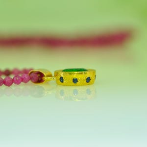 Pu&ograve; includere: Un pendente dorato con una pietra centrale verde e piccoli accenti blu, attaccato a una collana di perline rosa. Il pendente ha una superficie strutturata. Lo sfondo &egrave; verde chiaro.