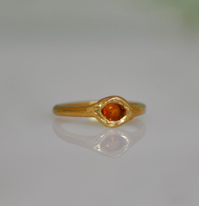 May include: A gold ring with an oval-shaped orange gemstone. The ring has a textured band and a bezel setting that holds the gemstone. The ring is displayed on a reflective surface.