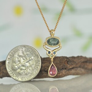 May include: A gold pendant necklace with a teardrop-shaped pink gemstone, a green oval gemstone, and two smaller blue gemstones. The pendant is hanging from a gold chain.