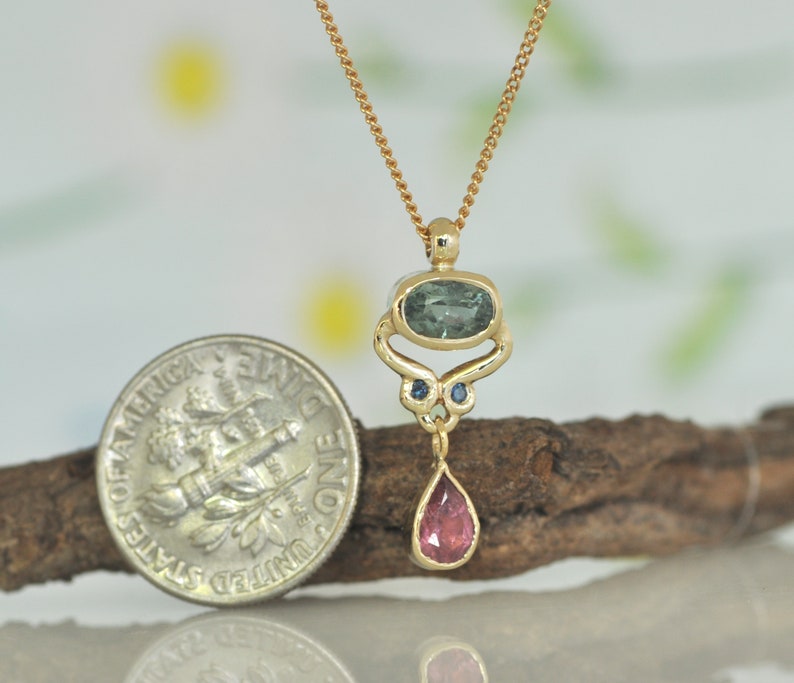 May include: A gold pendant necklace with a teardrop-shaped pink gemstone, a green oval gemstone, and two smaller blue gemstones. The pendant is hanging from a gold chain.