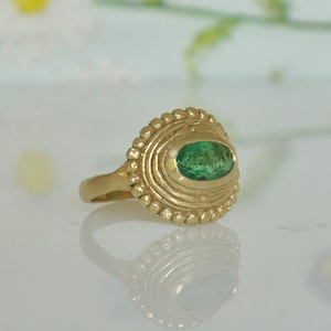 May include: A gold ring with a green emerald gemstone set in a textured, circular bezel. The ring has a simple band.