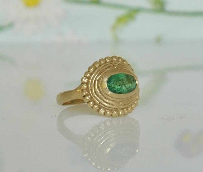 May include: A gold ring with a green emerald gemstone set in a textured, circular bezel. The ring has a simple band.