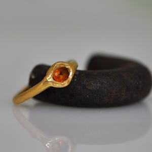 May include: A gold ring with an oval, orange gemstone. The ring is set in a textured gold band. The ring is resting on a dark, textured object, with a reflective surface below.