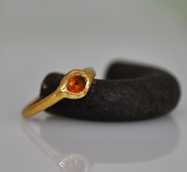 May include: A gold ring with an oval, orange gemstone. The ring is set in a textured gold band. The ring is resting on a dark, textured object, with a reflective surface below.