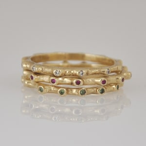 May include: Three gold rings stacked on top of each other. The top ring features small clear stones, the middle ring has red stones, and the bottom ring has green stones. The rings have a textured surface and are on a reflective surface.