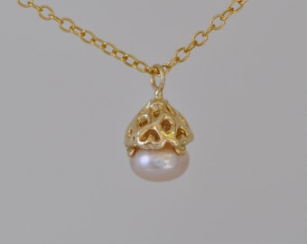 Könnte beinhalten: Eine zarte goldene Halskette mit einem herzförmigen Käfig-Anhänger, der eine einzelne Perle umschließt. Die Perle hat eine zarte rosafarbene Farbe.