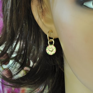 May include: Gold hoop earrings with a small charm dangling from the bottom. The charm is a round disc with a heart in the center and a border of green and pink gemstones.