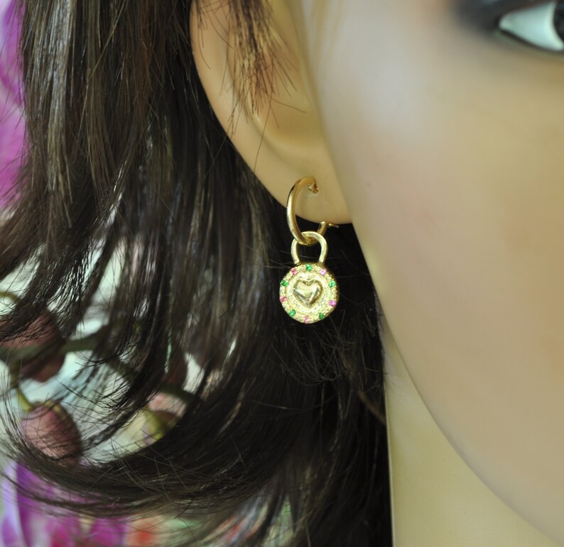May include: Gold hoop earrings with a small charm dangling from the bottom. The charm is a round disc with a heart in the center and a border of green and pink gemstones.