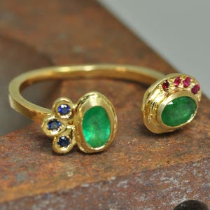 May include: A gold open-style ring featuring two oval-cut green emeralds as focal points. One side is accented with three small round blue sapphires, while the other side is adorned with three small round red rubies set in a textured gold base.