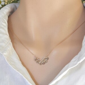 Puede incluir: Un collar de oro con un dise&ntilde;o delicado e intrincado que presenta una serie de remolinos conectados. El collar lo lleva una persona con una camisa blanca.