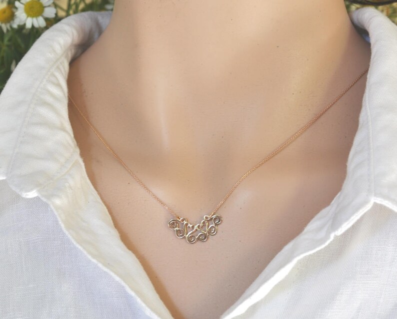 Puede incluir: Un collar de oro con un dise&ntilde;o delicado e intrincado que presenta una serie de remolinos conectados. El collar lo lleva una persona con una camisa blanca.