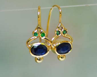 Könnte beinhalten: Ein Paar goldfarbene Ohrringe mit Haken-Design. Jeder Ohrring hat einen dunkelblauen, ovalen Stein und zwei kleine grüne Steine. Die Ohrringe haben ein dekoratives, verziertes Design.