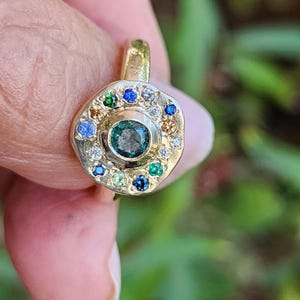 K&ouml;nnte beinhalten: Ein goldener Ring mit einem gr&uuml;nen Edelstein, der von einem Heiligenschein aus kleineren Edelsteinen in verschiedenen Farben, darunter Blau, Gr&uuml;n und Wei&szlig;, umgeben ist.