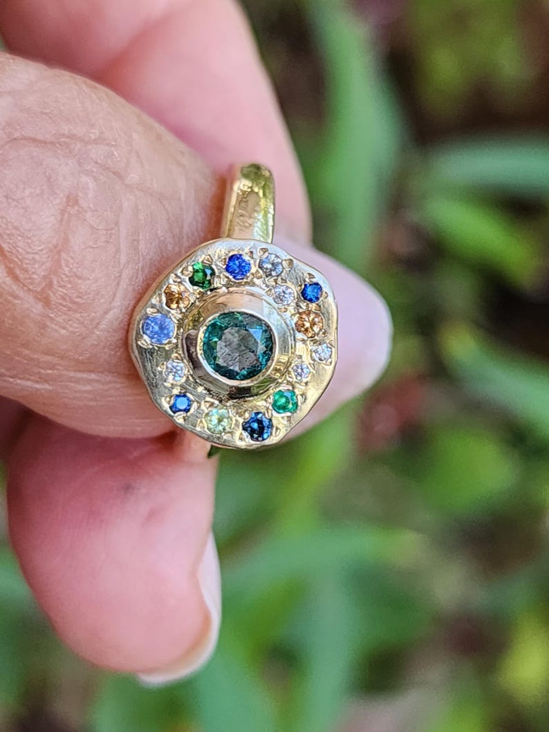 K&ouml;nnte beinhalten: Ein goldener Ring mit einem gr&uuml;nen Edelstein, der von einem Heiligenschein aus kleineren Edelsteinen in verschiedenen Farben, darunter Blau, Gr&uuml;n und Wei&szlig;, umgeben ist.