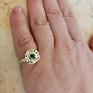 K&ouml;nnte beinhalten: Ein goldener Ring mit einem gr&uuml;nen Edelstein, der von kleineren bunten Edelsteinen umgeben ist.