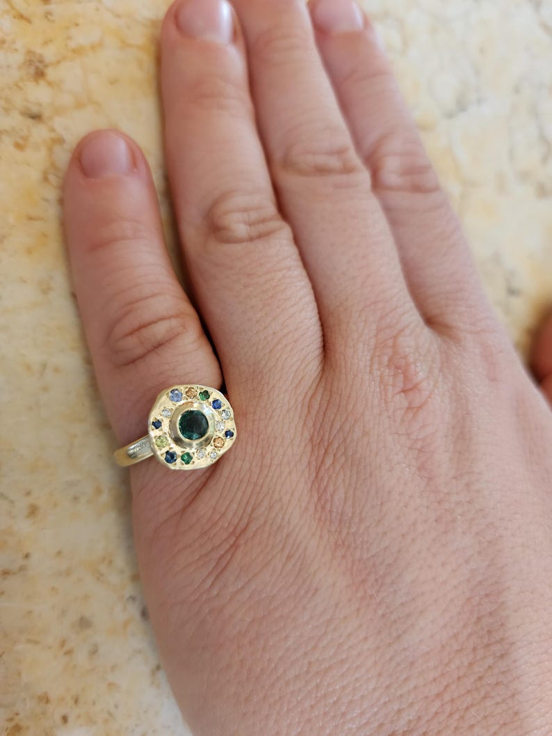 K&ouml;nnte beinhalten: Ein goldener Ring mit einem gr&uuml;nen Edelstein, der von kleineren bunten Edelsteinen umgeben ist.