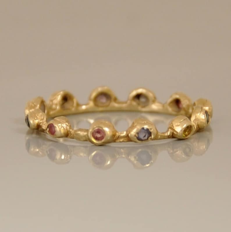 May include: A gold band ring with a textured surface and a row of colorful gemstones. The gemstones are set in a bezel setting and are arranged in a staggered pattern.