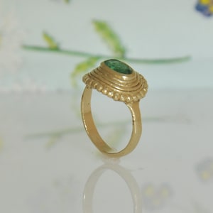 May include: A gold ring with a green gemstone set in a textured bezel. The ring has a simple band and a detailed setting.
