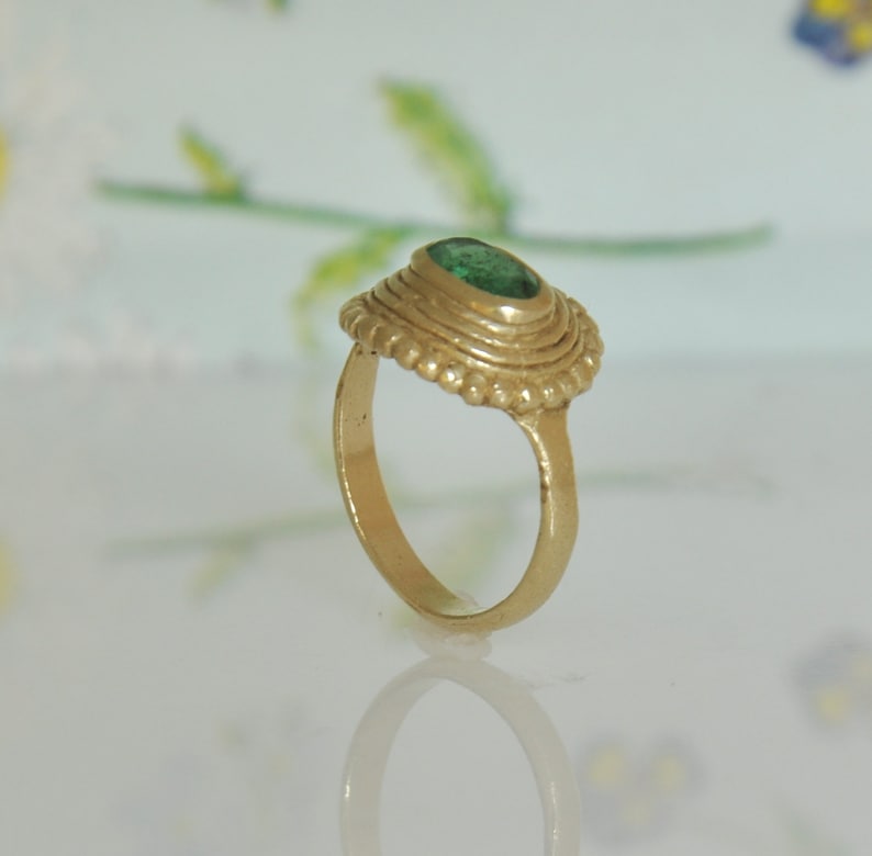 May include: A gold ring with a green gemstone set in a textured bezel. The ring has a simple band and a detailed setting.