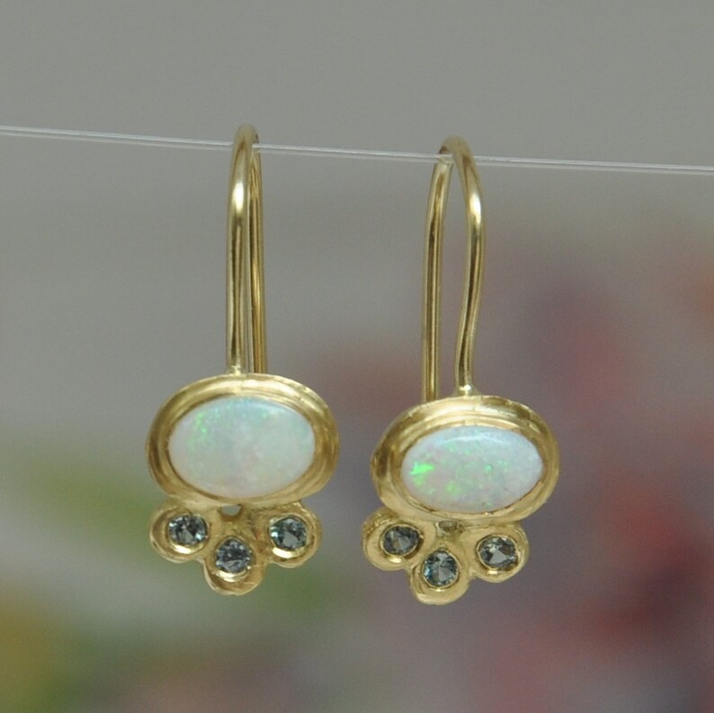 Op de afbeelding: Een paar gouden oorbellen met een witte opaal en kleine blauwe edelstenen. De oorbellen hebben een eenvoudig, elegant ontwerp.